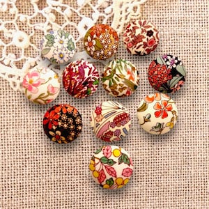 Peut inclure: Un ensemble de dix boutons ronds recouverts de tissu, chacun présentant un motif floral distinct avec des couleurs vives incluant le rose, l'orange, le rouge, le vert et le jaune sur des fonds blancs, noirs et crème, disposés en groupe.