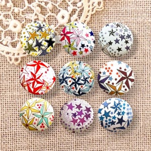 Peut inclure: Neuf boutons ronds recouverts de tissu disposés en une grille de trois par trois. Chaque bouton présente un motif unique d'étoiles de différentes couleurs, dont le rouge, le jaune, le bleu, le violet et le noir, sur fond de tissu blanc.