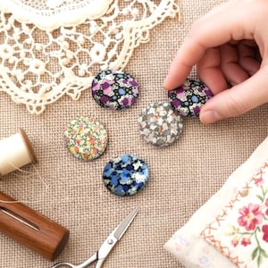 Pode incluir: Botões revestidos de tecido com estampas florais em tons de roxo, azul e amarelo, sobre tecido bege. Ferramentas de costura, renda e o texto "Liberty Folie" também estão presentes. Tema de artesanato ou costura.
