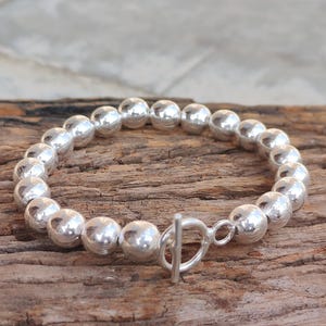 Op de afbeelding: Een zilveren kralenarmband met een tuimelsluiting. De armband heeft meerdere ronde, glanzende zilveren kralen die aan elkaar zijn geregen. De tuimelsluiting is een eenvoudig T-bar ontwerp. De armband wordt getoond op een houten oppervlak.