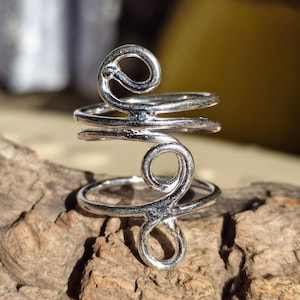 Könnte beinhalten: Ein silberfarbener Ring mit einem einzigartigen, wirbelnden Design. Der Ring hat zwei spiralförmige Akzente, die durch ein Band verbunden sind und einen offenen, verstellbaren Stil bilden. Der Ring wird auf einem Stück strukturiertem Holz präsentiert.
