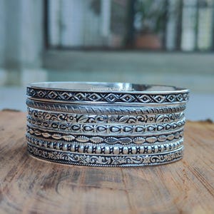 May include: A stack of seven silver bangles featuring diverse embossed patterns including geometric shapes, floral motifs, twisted rope textures, and beaded details. Each bangle has an oxidized finish that highlights the intricate metalwork designs.