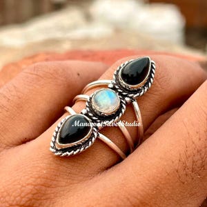 May include: A silver ring featuring a central round iridescent moonstone flanked by two black teardrop-shaped stones. The metal band has a twisted rope texture and splits into three distinct sections. The text ManavatSilverStudio is visible.