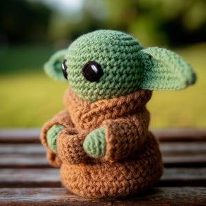 Pode incluir: Boneca de croch&ecirc; feita &agrave; m&atilde;o, &agrave; imagem de uma personagem popular. A boneca tem uma cabe&ccedil;a verde com olhos pretos grandes e orelhas pontudas. Ela est&aacute; vestindo uma t&uacute;nica marrom com m&atilde;os verdes. A boneca tem cerca de 15 cm de altura.