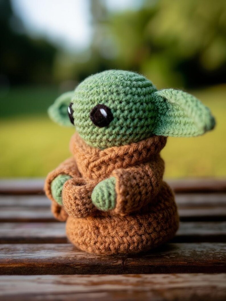 Pode incluir: Boneca de croch&ecirc; feita &agrave; m&atilde;o, &agrave; imagem de uma personagem popular. A boneca tem uma cabe&ccedil;a verde com olhos pretos grandes e orelhas pontudas. Ela est&aacute; vestindo uma t&uacute;nica marrom com m&atilde;os verdes. A boneca tem cerca de 15 cm de altura.
