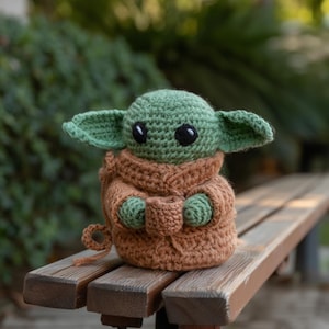 Pode incluir: Boneca de croch&ecirc; de um personagem com orelhas grandes, pele verde e olhos pretos. A boneca est&aacute; vestindo uma t&uacute;nica marrom e est&aacute; sentada em um banco de madeira. O fundo est&aacute; desfocado com folhagem verde.