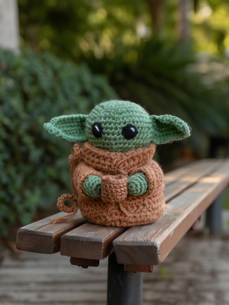 Pode incluir: Boneca de croch&ecirc; de um personagem com orelhas grandes, pele verde e olhos pretos. A boneca est&aacute; vestindo uma t&uacute;nica marrom e est&aacute; sentada em um banco de madeira. O fundo est&aacute; desfocado com folhagem verde.