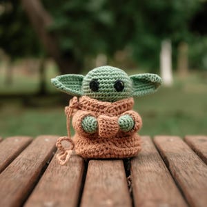 Pode incluir: Uma figura de croch&ecirc; de um personagem com orelhas grandes e uma t&uacute;nica marrom. A figura tem pele verde e olhos pretos grandes. A figura est&aacute; sentada em um banco de madeira. O personagem &eacute; de uma popular franquia de fic&ccedil;&atilde;o cient&iacute;fica.