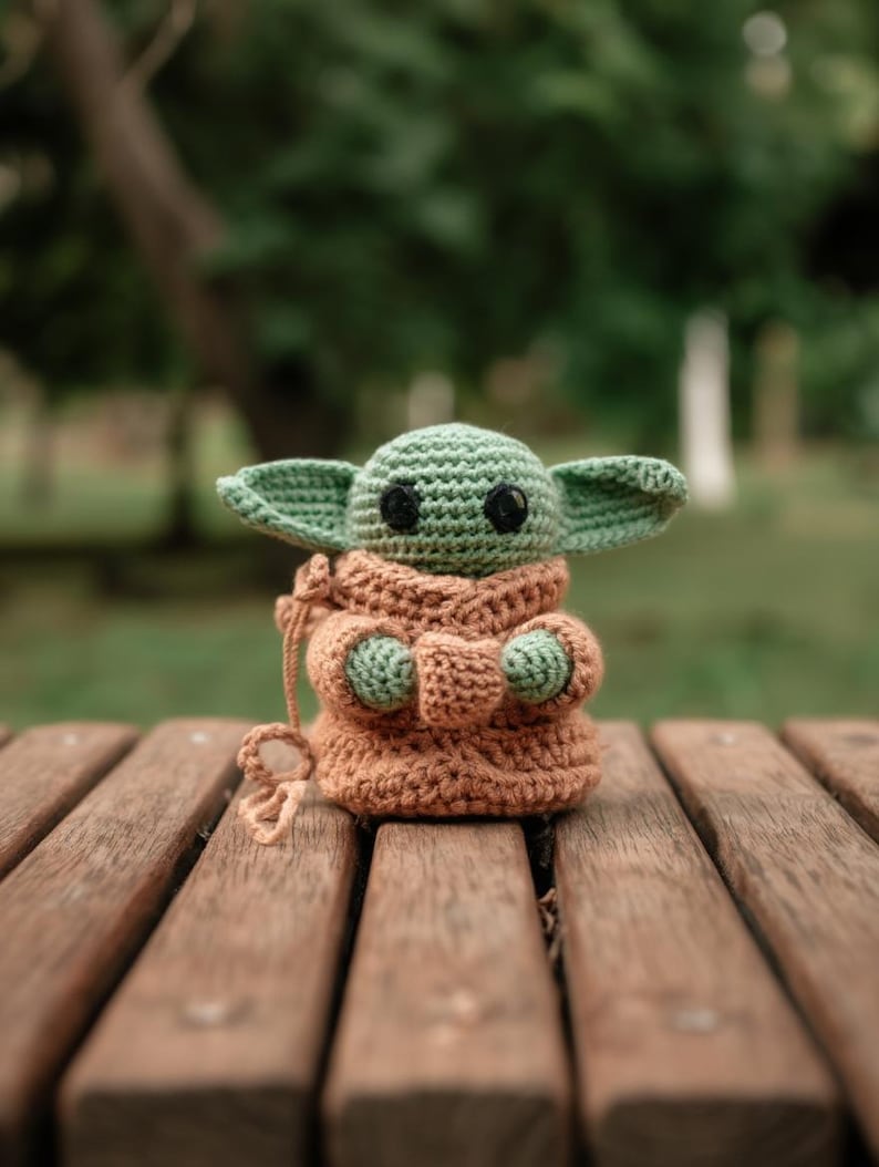 Pode incluir: Uma figura de croch&ecirc; de um personagem com orelhas grandes e uma t&uacute;nica marrom. A figura tem pele verde e olhos pretos grandes. A figura est&aacute; sentada em um banco de madeira. O personagem &eacute; de uma popular franquia de fic&ccedil;&atilde;o cient&iacute;fica.