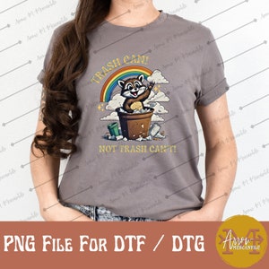 Pode incluir: Uma camiseta cinza com um gráfico de um guaxinim em um lixo. O guaxinim está sorrindo e segurando um pedaço de lixo. O texto "Trash Can! Not Trash Can't!" está impresso na camiseta.