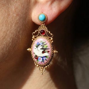 May include: Ornate gold-coloured drop earring with turquoise and red gemstone details. The oval pendant displays a painted bird and floral design on a pink background. The earring is highly detailed.