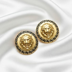 Puede incluir: Dos botones redondos dorados con una cabeza de león en el centro y un borde negro con diseño de greca. Los botones están sobre una tela blanca y sedosa, creando una estética lujosa. La cabeza de león es muy detallada.
