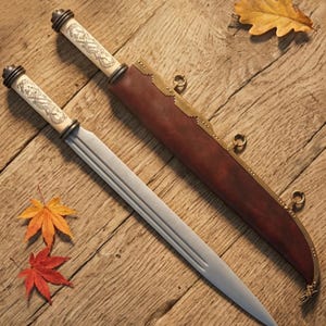 May include: Two decorative daggers with ornate ivory handles and silver blades rest on a wooden surface. A brown leather sheath with brass accents lies nearby. Autumn leaves in red, orange, and yellow are scattered around the daggers.