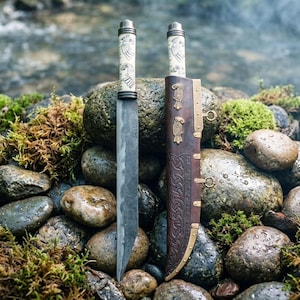 May include: Two handcrafted knives with ornate handles and blades, displayed with a leather sheath. The handles feature detailed carvings, and the sheath has decorative metal accents. The knives are set against a backdrop of moss-covered rocks and a flowing stream.