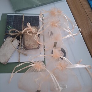 May include: Several small, sheer peach-colored organza bags with satin ribbon ties are arranged on a white surface. A few bags contain small metal charms. A bundle of cards tied with twine and a blank tag are also visible.