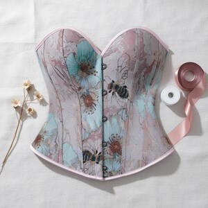 May include: A floral-patterned corset with a light pink trim and a row of black buttons down the front. The corset features a design of blue and pink flowers and bees. A spool of pink ribbon and a sprig of dried flowers are also visible.