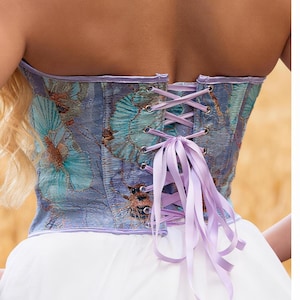 May include: A strapless corset with a butterfly pattern in shades of blue and purple. The corset has a lace-up back with a lavender ribbon. The bottom of the corset is paired with a white skirt.