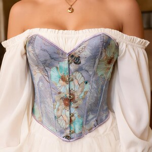 May include: A floral patterned corset in shades of blue, green, and purple. The corset features a sweetheart neckline and is adorned with metal clasps. It is worn with an off-the-shoulder white blouse.