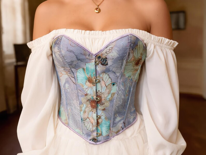 May include: A floral patterned corset in shades of blue, green, and purple. The corset features a sweetheart neckline and is adorned with metal clasps. It is worn with an off-the-shoulder white blouse.