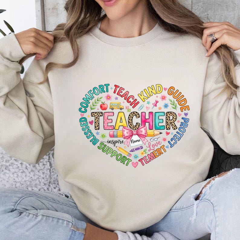 May include: Beige sweatshirt with a heart-shaped graphic for teachers. The design includes the word "TEACHER" in colorful letters, surrounded by words like "Comfort," "Kind," and "Support." A pencil, bow, and school bus are also featured.