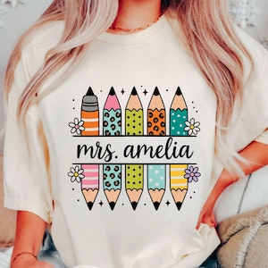 May include: Cream-colored t-shirt with a colorful pencil design. The pencils have various patterns and colors, including stripes, leopard print, and polka dots. The text "mrs. amelia" is displayed in a black banner.