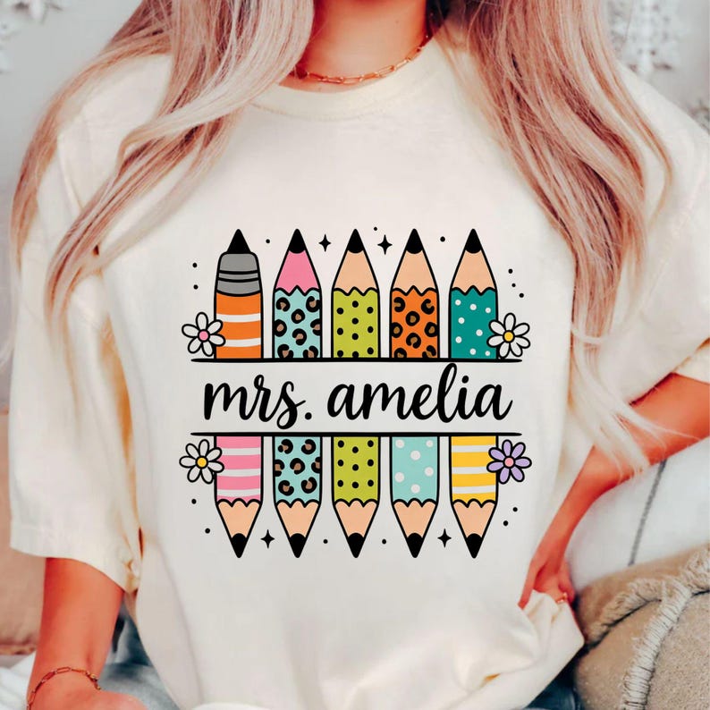 May include: Cream-colored t-shirt with a colorful pencil design. The pencils have various patterns and colors, including stripes, leopard print, and polka dots. The text "mrs. amelia" is displayed in a black banner.