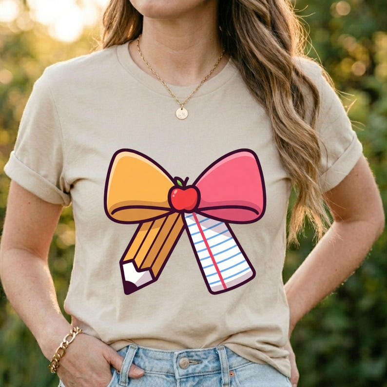 May include: Beige t-shirt featuring a colorful graphic of a bow made of a yellow pencil, pink and yellow bow, and a notepad. A red apple is in the center of the bow. The shirt is a casual style.