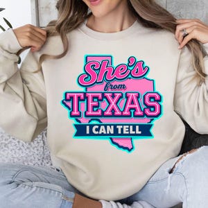 Puede incluir: Sudadera beige con el texto "She's from TEXAS I CAN TELL" en letras rosas y turquesas. La palabra "TEXAS" est&aacute; en letras grandes, con un contorno rosa del estado de Texas detr&aacute;s.