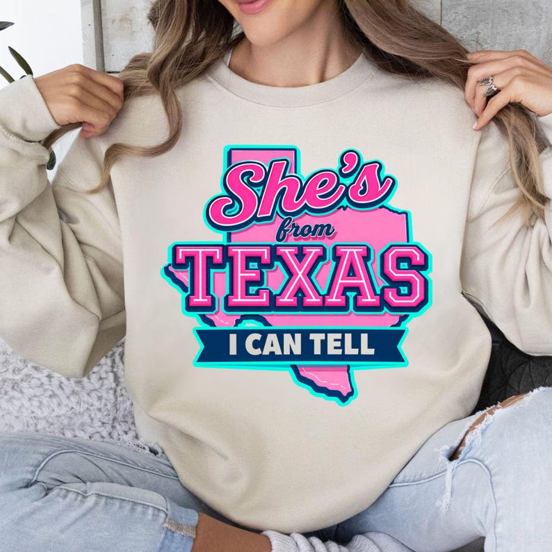 Puede incluir: Sudadera beige con el texto "She's from TEXAS I CAN TELL" en letras rosas y turquesas. La palabra "TEXAS" est&aacute; en letras grandes, con un contorno rosa del estado de Texas detr&aacute;s.