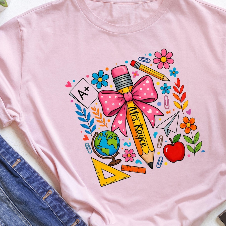 May include: Pink t-shirt featuring a colorful graphic design. The design includes a large yellow pencil with a pink bow, the text "Mrs. Kaycee", and various school-themed elements like flowers, a globe, and an apple.