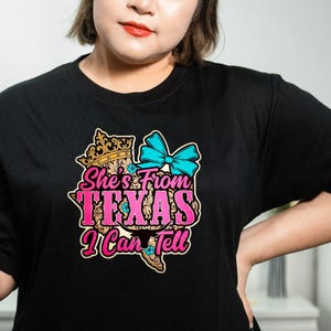 Puede incluir: Camiseta negra con un gr&aacute;fico colorido que dice "She's From Texas I Can Tell". El dise&ntilde;o incluye una corona, un lazo azul y el contorno de Texas en rosa y amarillo. Ropa casual.