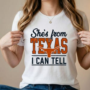 Puede incluir: Camiseta blanca con el texto "She's from TEXAS I CAN TELL" en azul marino y naranja. La palabra "TEXAS" presenta un gr&aacute;fico de cuerno largo. La camiseta es de cuello redondo y est&aacute; hecha de un material suave.