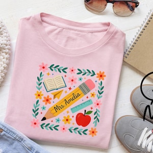 May include: A light pink t-shirt featuring a colorful graphic design. The design includes a yellow pencil with the text "Mrs. Amelia", an open book, a paper airplane, a ruler, an apple, and flowers. The shirt is a crew neck.