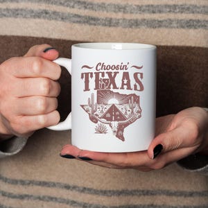 Puede incluir: Taza de cer&aacute;mica blanca con el texto "Choosin' TEXAS" en marr&oacute;n, con un estilo vintage. El dise&ntilde;o incluye un contorno de Texas con una carretera, cactus y una calavera de cuernos largos. La taza tiene un asa blanca.