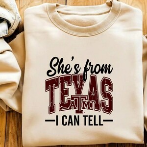 Puede incluir: Sudadera beige con cuello redondo y mangas largas. La parte delantera presenta el texto "She's from TEXAS A&M - I CAN TELL" en letras negras y granates. La sudadera est&aacute; hecha de un material suave.