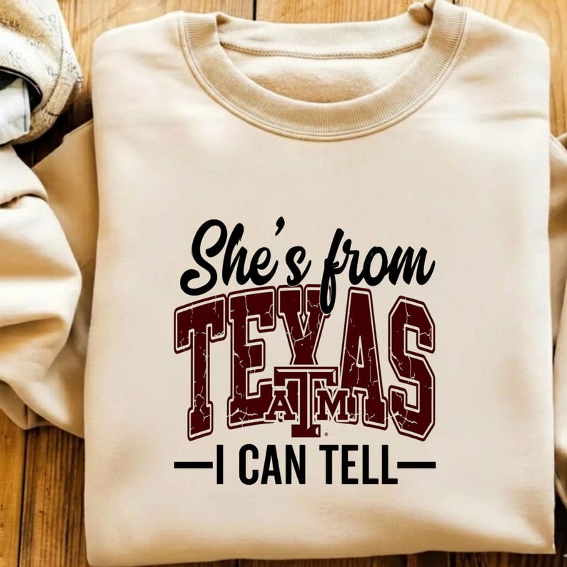 Puede incluir: Sudadera beige con cuello redondo y mangas largas. La parte delantera presenta el texto "She's from TEXAS A&M - I CAN TELL" en letras negras y granates. La sudadera est&aacute; hecha de un material suave.