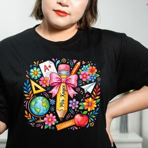 May include: Black t-shirt featuring a colorful school-themed graphic. The design includes a large pencil with a pink bow, the text "Mrs. Kayce", flowers, a globe, an apple, and a paper airplane. The shirt has a crew neck.