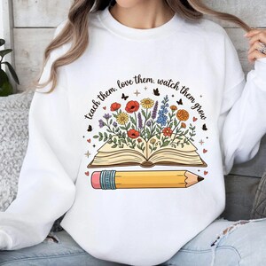 May include: White sweatshirt with a colorful graphic. The graphic features an open book with flowers and a pencil. The text reads "teach them, love them, watch them grow".