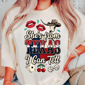 Puede incluir: Camiseta color crema con un dise&ntilde;o gr&aacute;fico que presenta labios rojos, una calavera de toro con sombrero de vaquero y el texto "She's from TEXAS I Can Tell". Gr&aacute;ficos adicionales incluyen naipes, cerezas y dados.