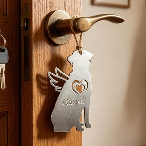 Puede incluir: Un colgador de puerta de metal plateado con forma de perro con alas de ángel y un corazón recortado, colgado de un pomo. El nombre "Caspar" está grabado en el perro. La puerta es de madera.