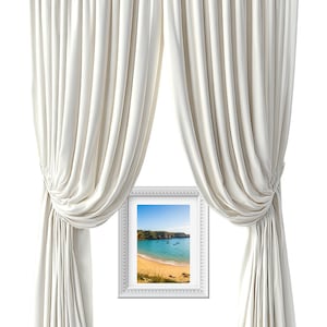 Puede incluir: Un par de cortinas de color blanco roto que llegan hasta el suelo con pliegues verticales, recogidas a los lados, enmarcando un marco de fotos rectangular blanco que contiene una fotografía de una playa, mar turquesa y acantilados.