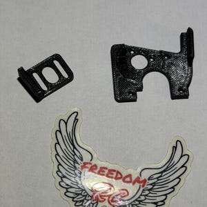 May include: Two black 3D-printed plastic components with textured surfaces and a sticker featuring white wings, red text reading FREEDOM, and red text reading RC on a white background. The components have cutouts and mounting holes.