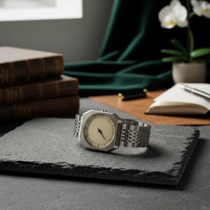 May include: A silver-toned wristwatch with a cream-colored face and a rectangular case. The watch has a linked metal band and is displayed on a dark gray slate surface. The watch is a classic, minimalist design.