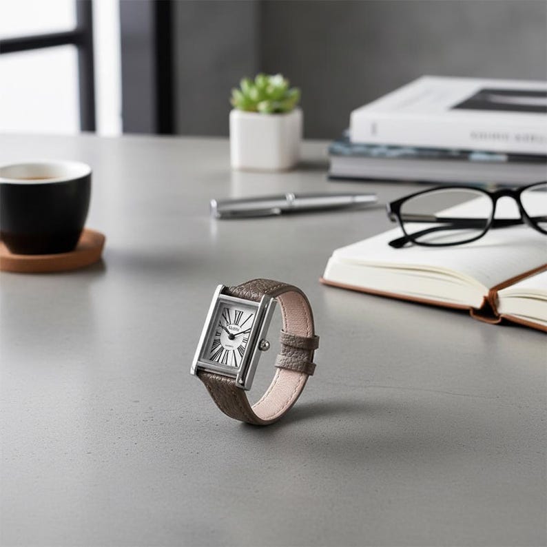 May include: A rectangular silver wristwatch with a brown leather band. The watch face is white with black Roman numerals and hands. The watch is displayed on a gray surface, with a cup of coffee, a pen, and a notebook in the background.