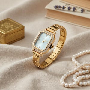May include: A gold wristwatch with a rectangular face and a light blue dial. The watch has a crystal-studded bezel and a matching gold band. A gold box, a book, and a pearl necklace are in the background.