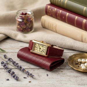 Könnte beinhalten: Eine goldene rechteckige Armbanduhr mit einem weinroten Lederarmband liegt auf einem passenden weinroten Lederportemonnaie. Das Zifferblatt der Uhr ist cremefarben mit schwarzen römischen Ziffern. Ein kleines Glas mit getrockneten Blumen und Bücher befinden sich im Hintergrund.