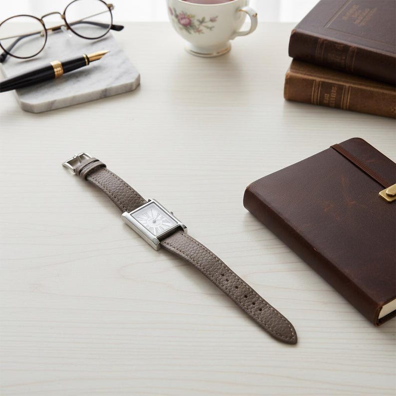 May include: A rectangular silver wristwatch with a gray leather band lies on a white surface. The watch face has a white background with a decorative design. Other items on the surface include a pen, glasses, a teacup, and a leather-bound notebook.
