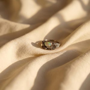 Könnte beinhalten: Ein silberner Ring mit einem ovalen, schillernden Opalstein in der Mitte. Die Ringschiene ist mit detaillierten Metallarbeiten versehen, darunter ein kleines kreisförmiges, fächerartiges Muster an der Seite der Fassung, das eine Textur erzeugt.