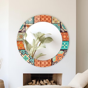 May include: A circular wall mirror featuring a wide frame decorated with a vibrant, multi-coloured tile pattern in shades of orange, teal, and cream. The reflective surface shows a clear view of a green plant with broad, pointed leaves.