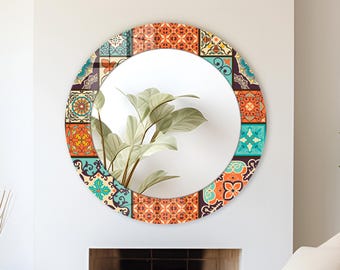 May include: A circular wall mirror featuring a wide frame decorated with a vibrant, multi-colored tile pattern in shades of orange, teal, and cream. The reflective surface shows a clear view of a green plant with broad, pointed leaves.